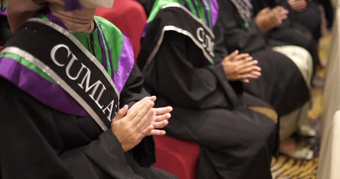 Close Up Happy graduates in hats and robes siting in row and applauding. Cheerful male and female university or college students having fun at graduation ceremony. Clapping hands
