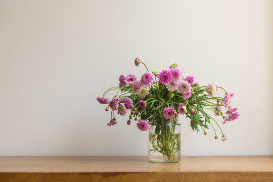 Close up of handpicked purple daisies in glass jar on oak table against beige wall (selective focus)