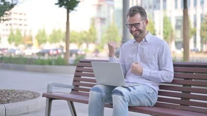Young Adult Man Celebrating Success on Laptop while Sitting Outdoor on Bench