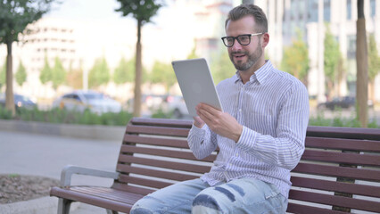 Fototapeta premium Young Adult Man Celebrating Online Win on Tablet while Sitting Outdoor on Bench