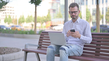 Young Adult Man Using Smartphone and Laptop while Sitting Outdoor on Bench