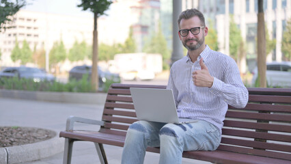 Young Adult Man with Laptop Showing Thumbs Up Sign While Sitting Outdoor on Bench
