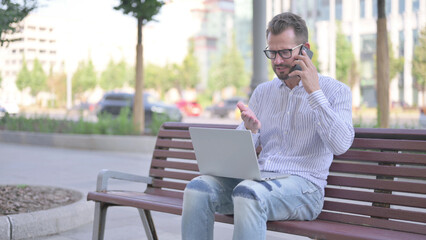Fototapeta premium Angry Young Adult Man Talking on Phone and using Laptop while Sitting Outdoor on Bench