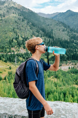 Young traveller with backpack stands on the backdrop of a stunning view with green mountains and drinks clear water from a blue bottle. High quality photo