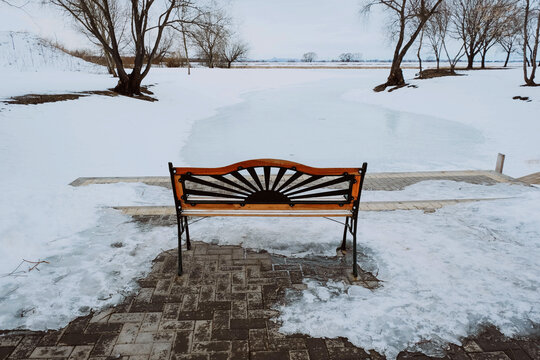 Cute bench near snow-covered river. Cozy winter walk in park. © Slepitssskaya