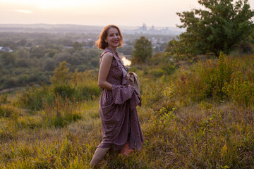 Beautiful young smiling girl in a long brown dress walks along the lawn. Happy woman walks at sunset on a hill overlooking the river. Concept of having rest in park during summer holidays or weekends