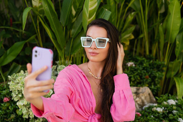 Young beautiful brunette in a pink shirt, neck jewelry, necklace, trendy sunglasses sits on a background of tropical leaves, takes a selfie on the phone