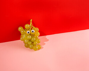 A bunch of grapes with googly eyes reposed on pink and red background from angle.