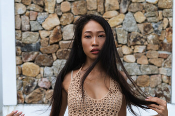 Tender sensitive Asian woman in a knitted dress and swimsuit posing near a brick wall, chic smooth long hair, romantic perfect clear skin