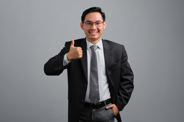 Portrait of smiling handsome young businessman in formal suit showing thumb up isolated on grey background