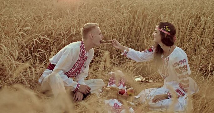 Man and a woman Ukrainians have lunch in the field, a towel is spread out, they feed each other. Man and a woman in beautiful national costumes, they are happy, lunch in a wheat field. 4k, ProRes