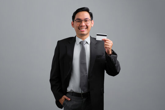 Portrait of smiling handsome young businessman in formal suit holding credit card isolated on grey background