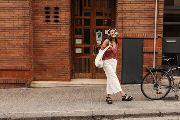 Full length happy young caucasian woman in headphones looking at camera standing outdoors. Brunette wears casual clothes in spring. Audio technology concept