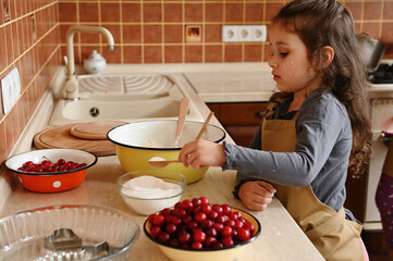 Beautiful child, little girl in chef apron holding a scoop with white sugar and adding into dry ingredients in the bowl with flour, kneading dough for a festive cake with cherries in the home kitchen