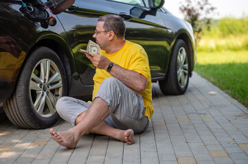 worried man sitting on the pavement next to a car near an open fuel tank, fuel price rise concept