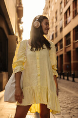 Nice young caucasian brunette woman with headphones spends leisure time in city sunny weather. Model wears yellow summer dress, bag. Lifestyle concept