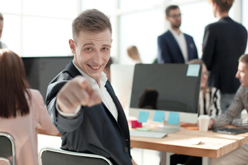 smiling young employee sitting at his Desk and pointing at you