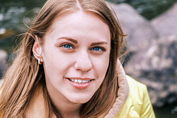 Portrait of young attractive blue-eyed woman looking at camera and smiling in yellow jacket outdoors.