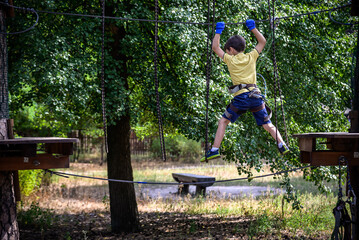Strong excited young boy playing outdoors in rope park. Caucasian child dressed in casual clothes and sneakers at warm sunny day. Active leisure time with children concept
