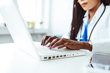 Close-up hands of unrecognizable female physician in medical uniform working typing on laptop keyboard sitting at desk on background of window in dark office room.