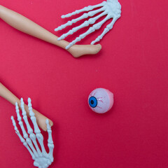 The hands of the skeleton hold the legs and a creepy eye watches it all, on a red background. Minimal flat lay scenes.