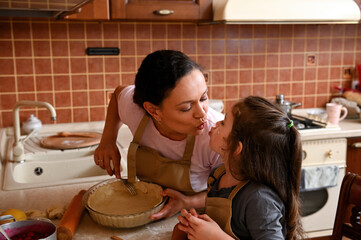 Beautiful dark-haired Hispanic young woman wearing beige chef apron, a loving mom kissing her cute...