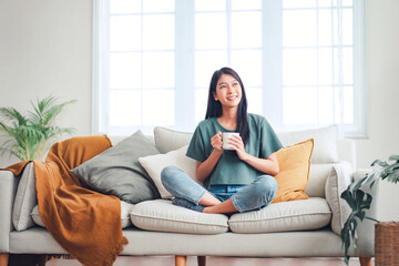 Happy young asian woman drinking coffee relaxing on sofa at home. Smiling female enjoying resting...