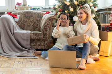 Indoor shot of beautiful happy young woman shopping online on laptop in cozy Christmas interior. Mother on the floor next the Christmas tree and sofa and daughter embrace her, shopping gifts.