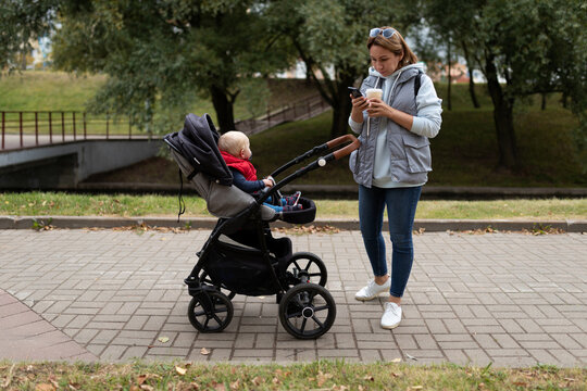 a young mother drinks coffee and watches a mobile phone while walking with a baby in a stroller around the city