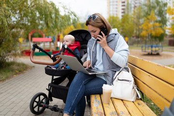 Fototapeta na wymiar a young mother sitting on a bench next to a stroller with a child works online on a laptop and answers a phone call