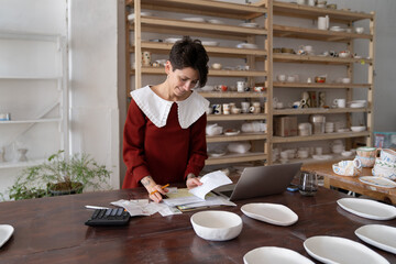 Smiling ceramic shop owner calculating business costs while working in pottery shop, female artisan making analysis of pottery studio expenses, doing bookeeping, working with invoices and receipts