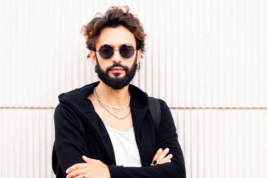 portrait of a beautiful young man with beard and sunglasses leaning in a white wall, concept of urban lifestyle and stylish clothing