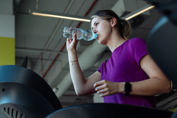 Tired overweight girl run on treadmill at fitness gym and quenching thirst drink water from bottle,...