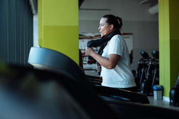 Adult fat woman energetically run breathing through mouth on treadmill at fitness gym, side view. Lady with towel around neck do cardio workout on training apparatus. Weight loss, sport lifestyle