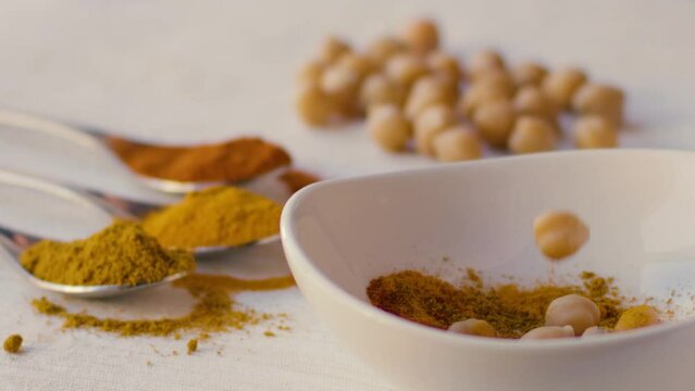 Chickpeas fall in slow motion into a white bowl filled with curry powder, other chickpeas and three spoons with curry powder, saffron and turmeric powder are also seen on the table