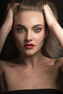 Studio portrait of beautiful blonde woman with long hair, green eyes and red lipstick looking to camera. Model holding hands over head and brushing hair with fingers. Toned image in beige color