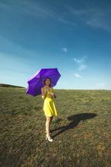 woman in yellow sundress and purple umbrella walking on the green meadow at day in summer