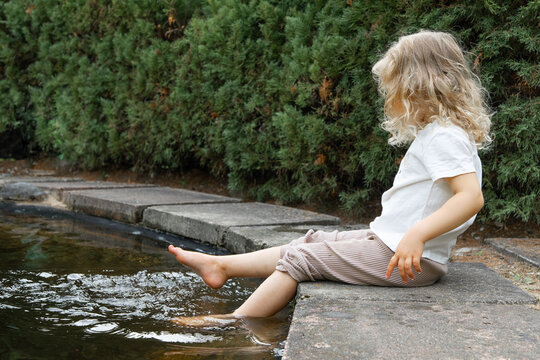 Unrecognizable little blonde curly barefoot girl washing legs, foot in lake water in natural park in hot summer outdoors