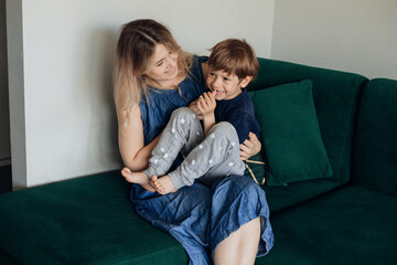 Blond smiling, cheerful and kind woman and boy, her son playing and embracing, tickling together at home. Child care - obrazy, fototapety, plakaty