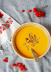 Yellow pumpkin cream soup squash with seeds, spoon in bowl, gray textile fabric background, embroidered ukrainian ornament towel, rowan bunches; autumn seasonal healthy vegetarian food, vertical