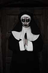 Portrait of scary devilish nun with clasped hands near wooden door outdoors. Halloween party look