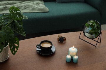 Wooden table with cup of coffee, candles, mirror, wristwatch and houseplant near sofa in room....