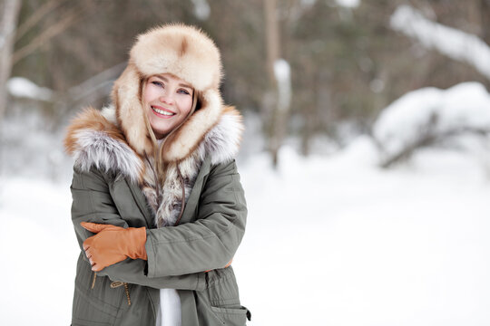 Portrait of beautiful happy girl standing in nature winter snow forest. Smiling woman wearing warm clothes, winter parka jacket, fur hat, leather gloves