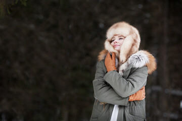 Woman wrap and hug herself on dark background, wearing winter jacket, fur ear flaps hat and leather glove. Cozy portrait