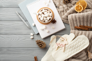 Top view photo of cup of hot drinking with marshmallow on rattan placemat planner pen reindeer ornament mittens scarf pine cones cinnamon sticks and dried orange slices on grey wooden table background