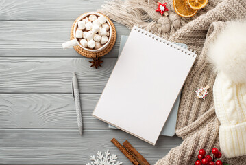 Top view photo of notepad pen bobble hat plaid cup of hot drinking with marshmallow serving mat snowflake cinnamon sticks viburnum and dried orange slices on grey wooden desk background with copyspace