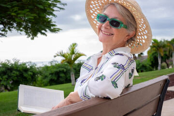 Portrait of cheerful senior woman with green sunglasses and hat sitting on a bench in public park reading  a book - caucasian lady looking at camera smiling
