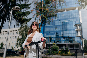 Naklejka na ściany i meble Beautiful young woman in sunglasses and white suit standing on her electric scooter near modern building and looking away