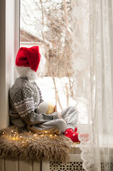 a child in festive clothes and a red Christmas hat is sitting by the window with a golden ball in his hand