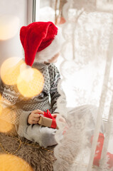 a child in festive clothes and a red Christmas hat is sitting by the window with a golden bokeh on the side